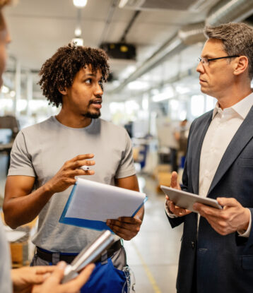 Mid adult engineer using touchpad and communicating with African American worker about product reports in industrial building.